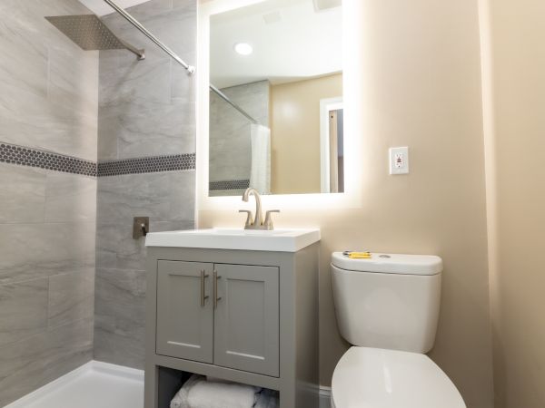 A modern bathroom with a shower, small sink with mirror above, gray cabinet, and a toilet on the right, all in neutral tones.