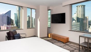 A modern hotel room with large windows, a bed, a chair, a TV, and a view of a cityscape with tall buildings outside.