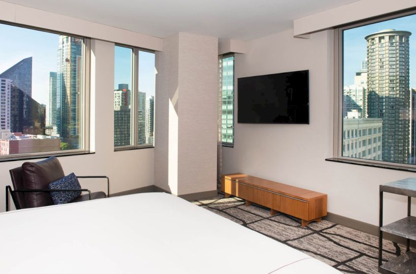 A modern hotel room with large windows, a bed, a chair, a TV, and a view of a cityscape with tall buildings outside.
