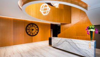 A modern lobby features a marble counter, large wall clock, wooden accents, spiral ceiling, and decorative light fixtures with flowers.
