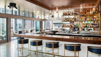 A sleek bar with blue stools, stocked shelves, modern lights, and large windows overlooking a cityscape.