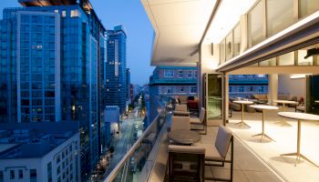 A modern cityscape at dusk with tall buildings and a terrace featuring tables and seating.