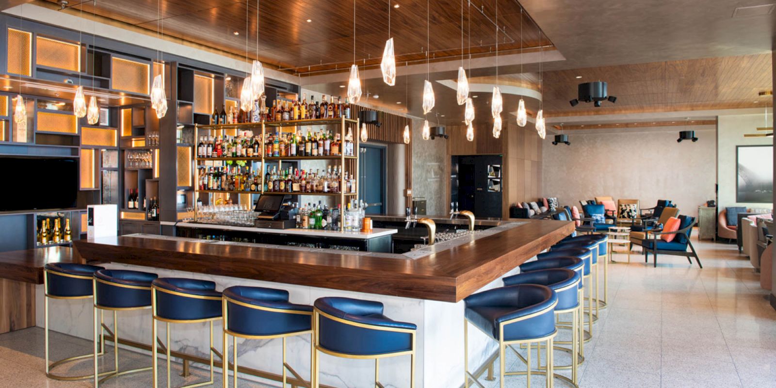 A modern bar with stylish blue and gold seating, a well-stocked back shelf, and pendant lighting creates a warm, inviting atmosphere.