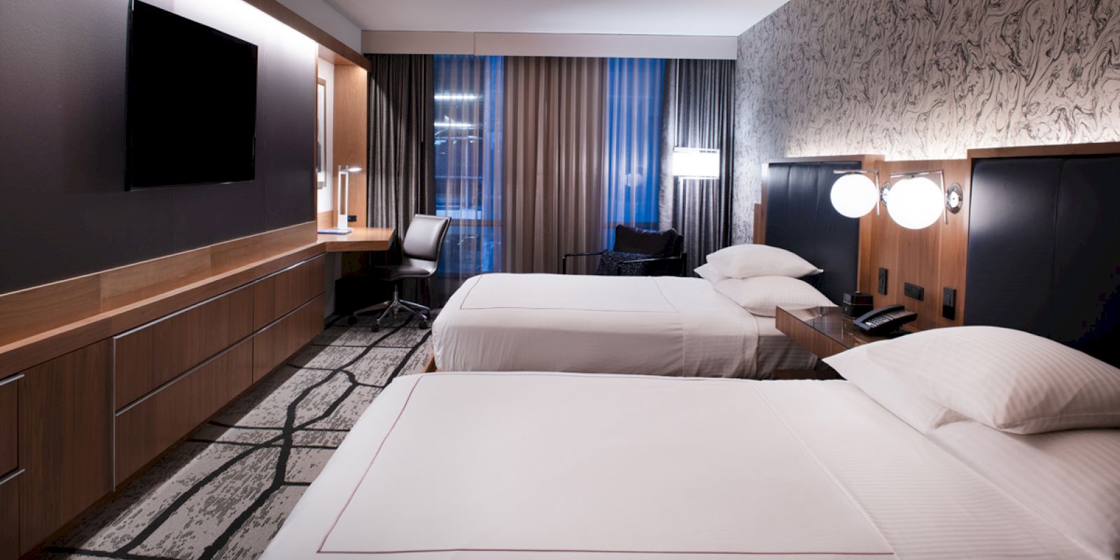 A modern hotel room with two beds, a TV, desk, and lamps, featuring a stylish decor and carpet design.