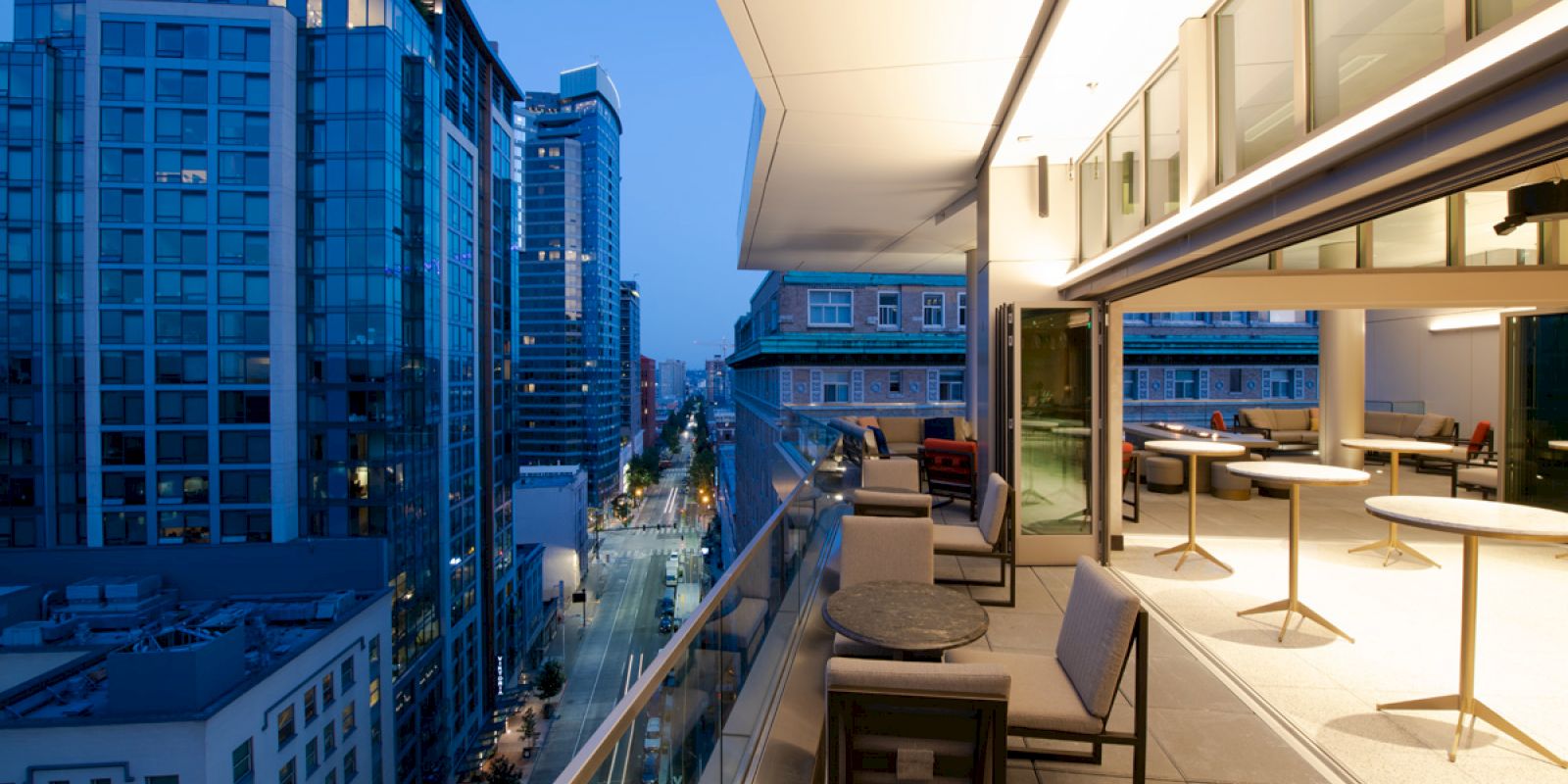 A modern rooftop terrace overlooks a cityscape with tall buildings at dusk, featuring seating and tables under soft lighting.
