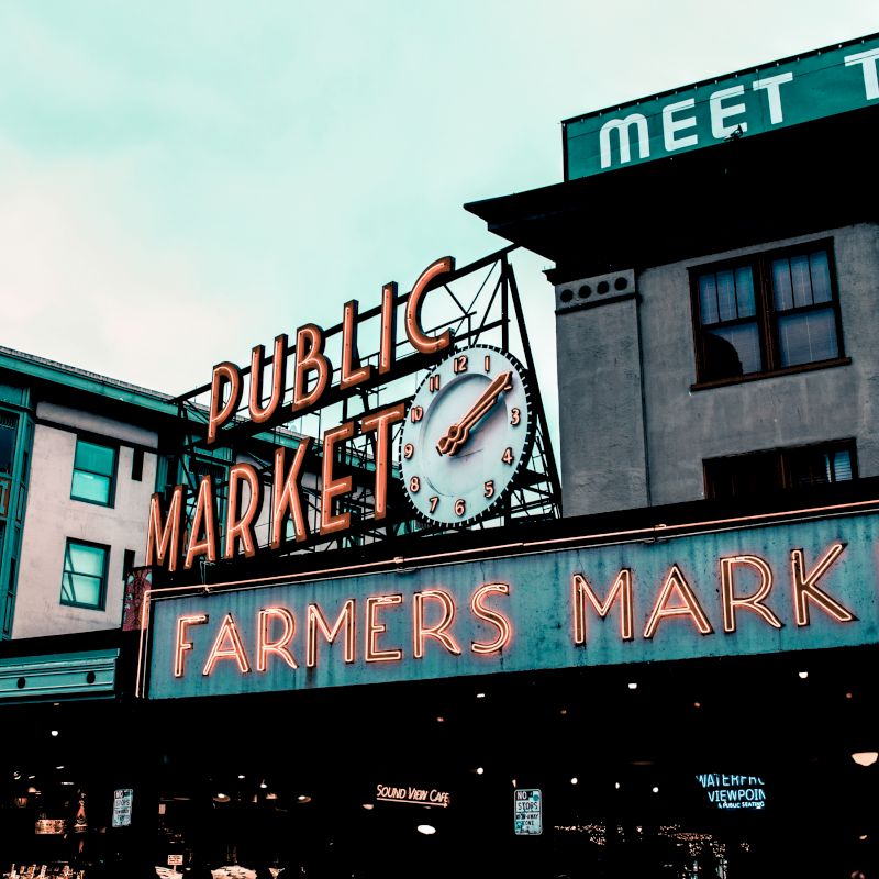 The image shows an iconic neon sign for a public market and farmers market, with a clock, against a backdrop of buildings.