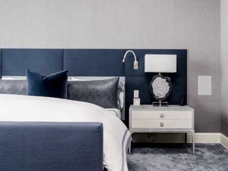 A modern bedroom features a navy bed, white linens, and a gray nightstand with a lamp.