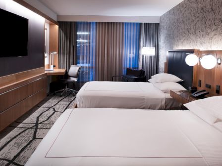 The image shows a modern hotel room with two beds, a TV, a desk, and stylish decor, lit by elegant lamps and natural light.