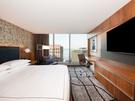 A modern hotel room with a large bed, TV, desk, and a window with city views. The room is stylish and well-lit.