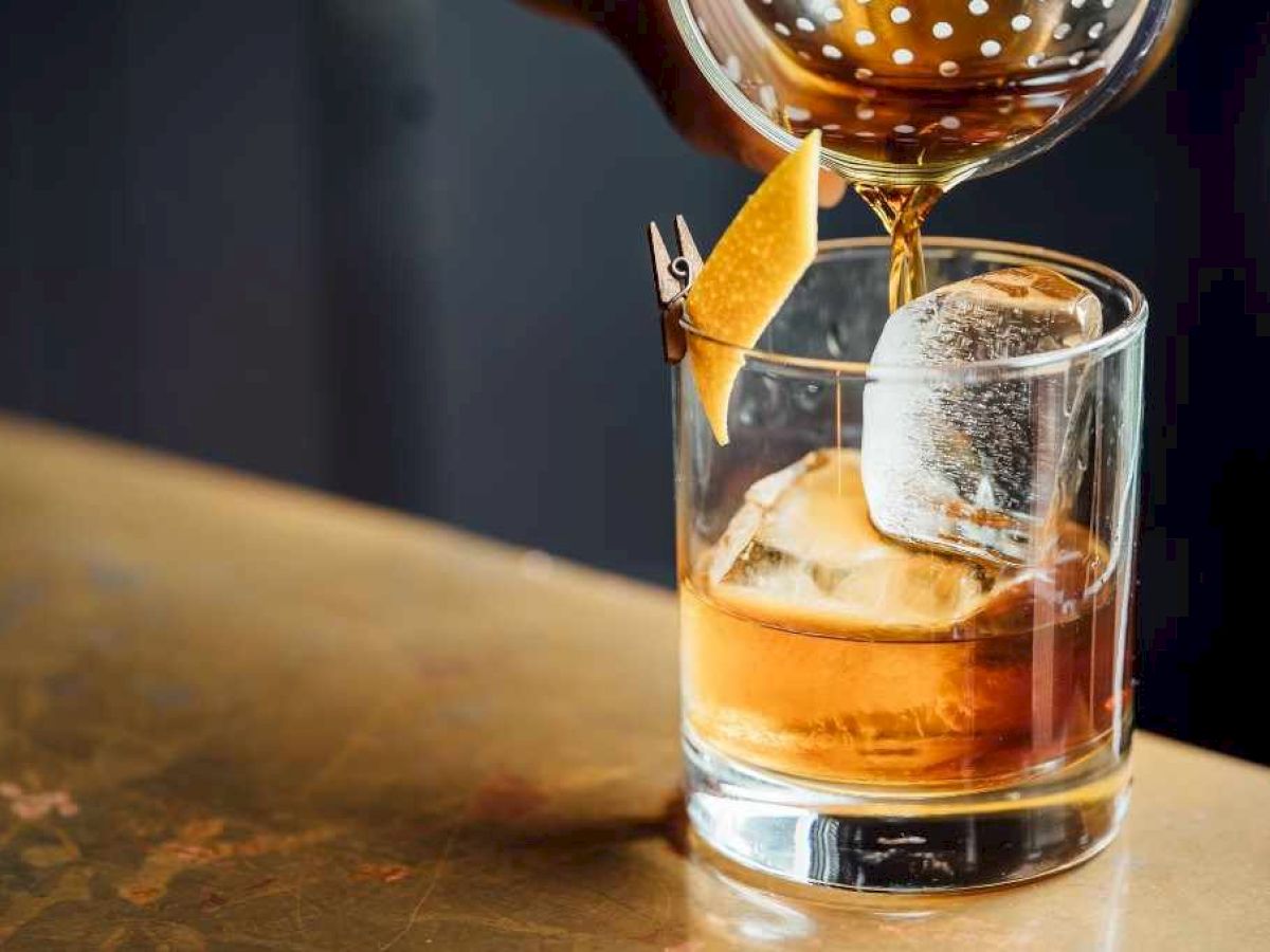 A cocktail is being poured over ice in a glass with a lemon twist garnish on a bar counter.
