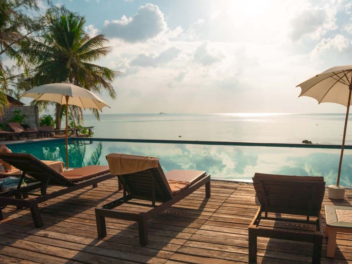 Lounge chairs and umbrellas by a pool with a scenic view of the ocean and palm trees under a partly cloudy sky.