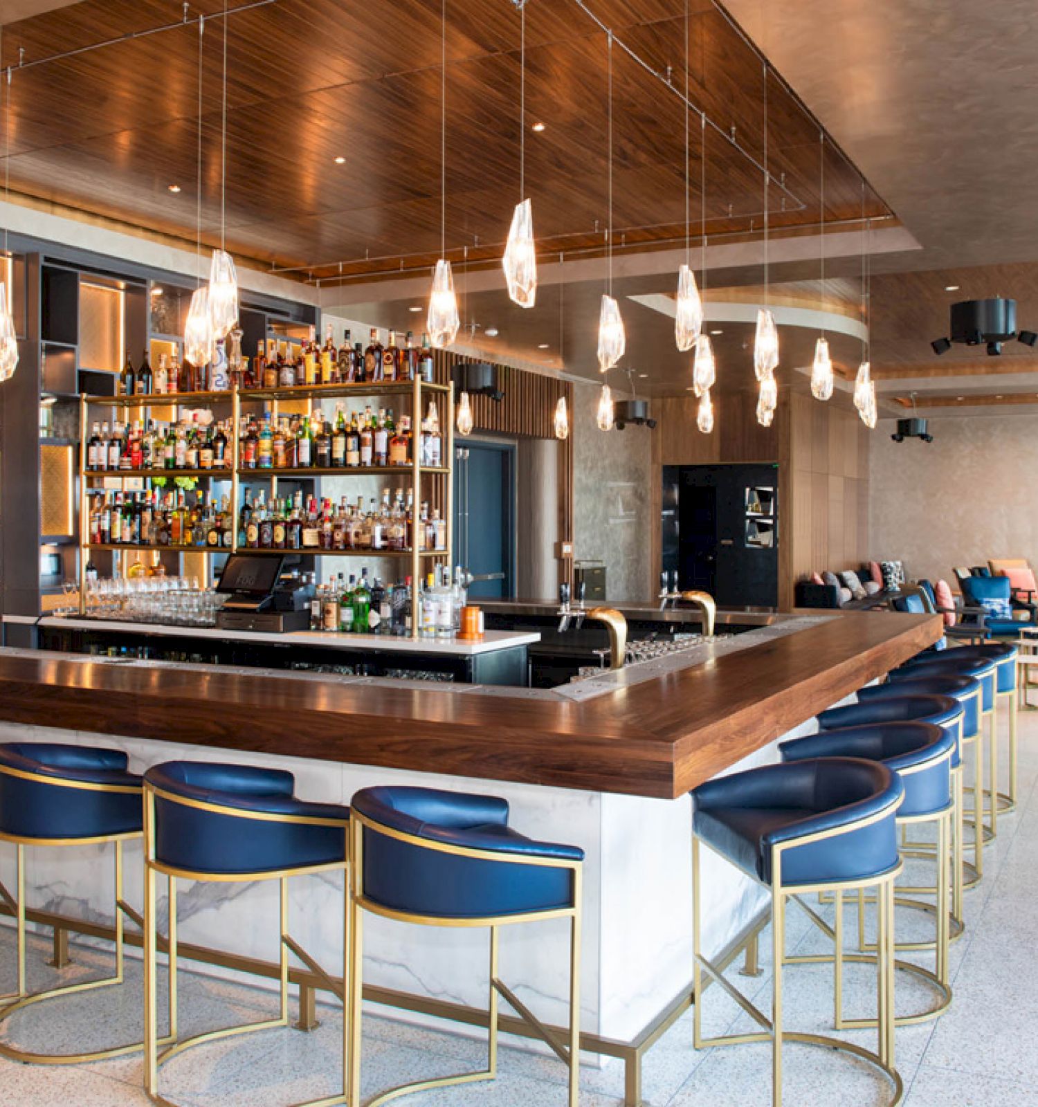 Stylish bar with modern seating, overhead lighting, and a fully stocked counter. Cozy lounge area with sofas in the background.
