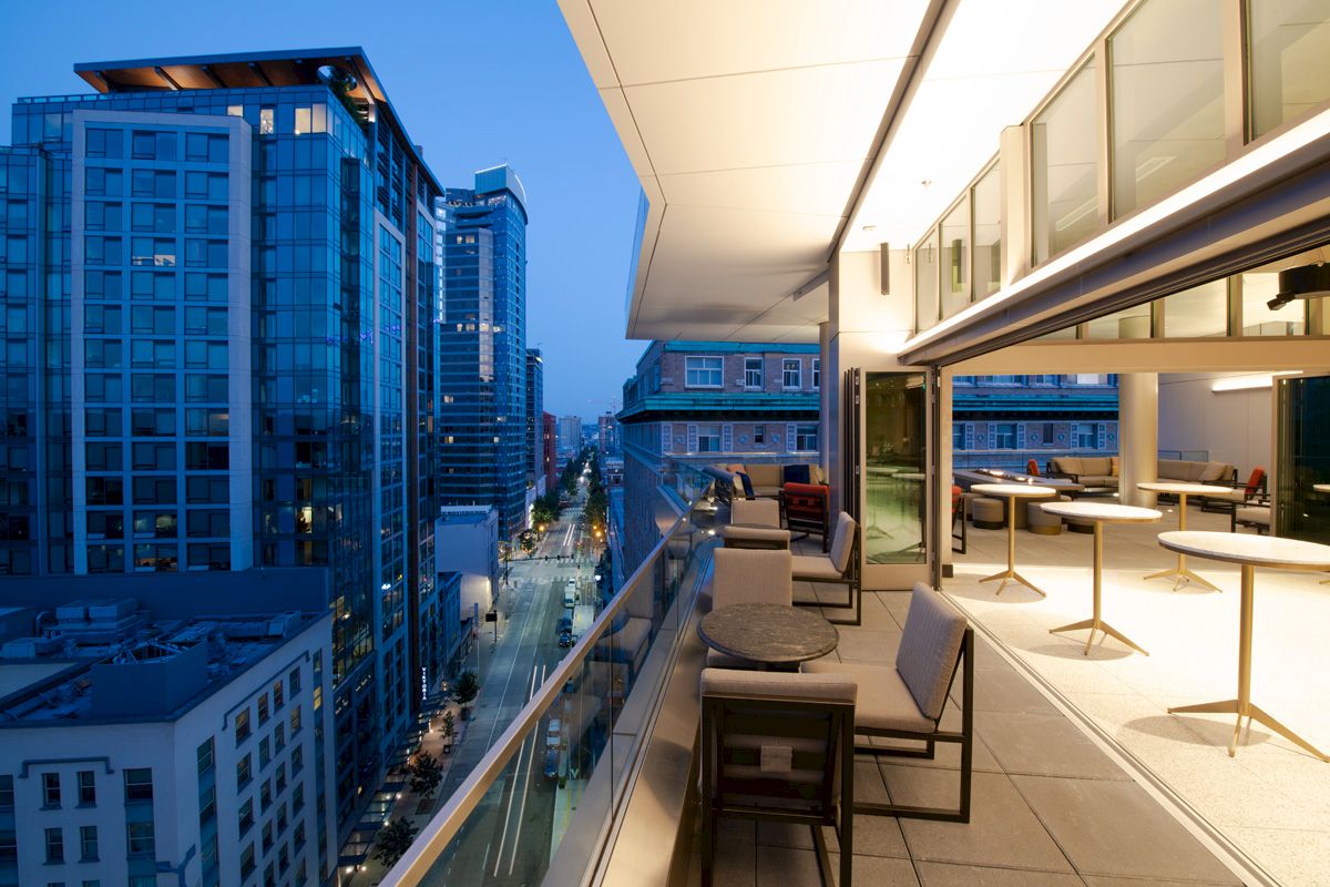A modern rooftop patio with seating overlooks a city street and adjacent buildings at dusk.