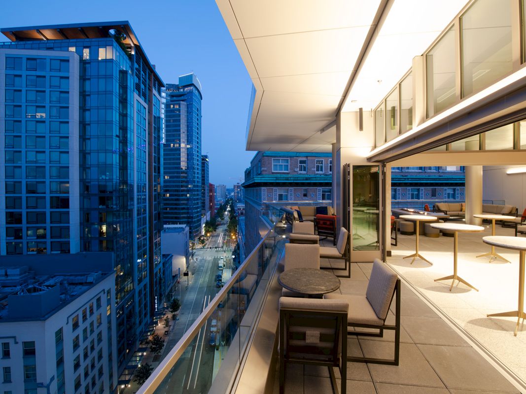 A modern rooftop patio with seating overlooks a city street and adjacent buildings at dusk.
