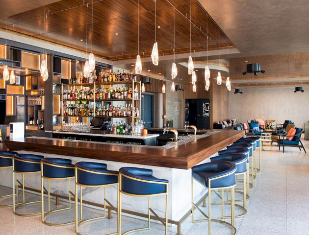 A modern bar with a wooden counter, blue barstools, hanging lights, and a well-stocked liquor shelf; lounge seating visible in the background.