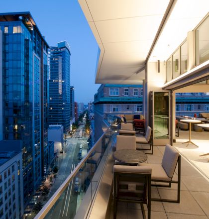 A modern rooftop terrace with tables and seating overlooks a cityscape of tall buildings and streets at dusk.