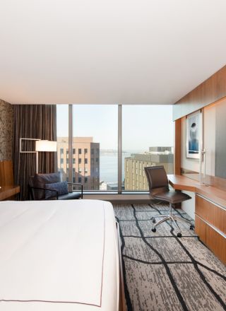 A modern hotel room with a large bed, desk, chair, wall-mounted TV, and a city view through large windows is shown.
