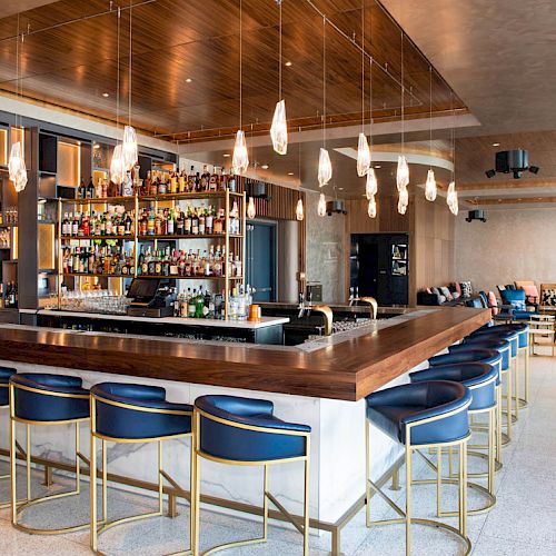A modern bar with blue stools, hanging lights, and a stocked drinks display, surrounded by cozy seating in a spacious, stylish setting.