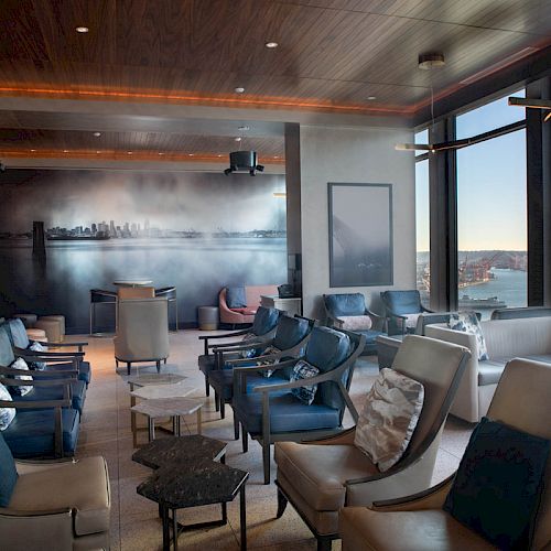 A modern lounge with leather chairs, small tables, large windows offering a scenic view, and a wall mural depicting a city skyline.