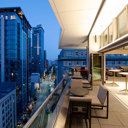 A modern cityscape view featuring tall buildings and a stylish balcony setup with tables and chairs at dusk.