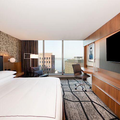 A modern hotel room features a large bed, wall art, a flat-screen TV, a desk with a chair, and a window overlooking city buildings.