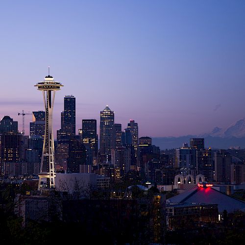 Seattle skyline at dusk with the Space Needle and Mount Rainier in the background, set against a purple and blue sky.