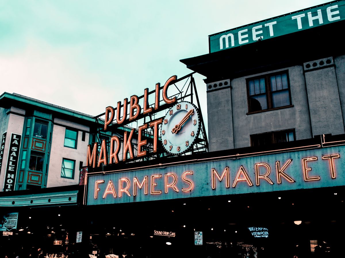 A public market sign with a clock and "Farmers Market" in neon lights on a building, under a cloudy sky.