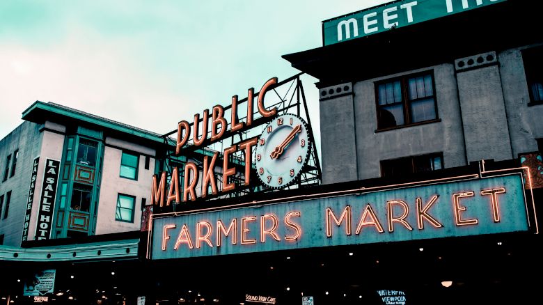 A public market sign with a clock and "Farmers Market" in neon lights on a building, under a cloudy sky.