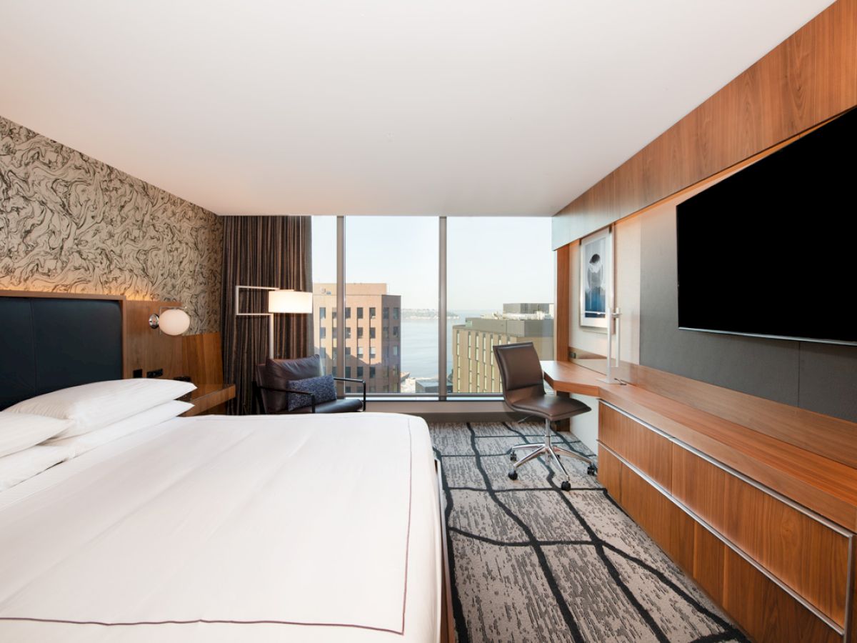 A modern hotel room with a large bed, wall-mounted TV, desk, chair, and a window view of buildings and the waterfront.