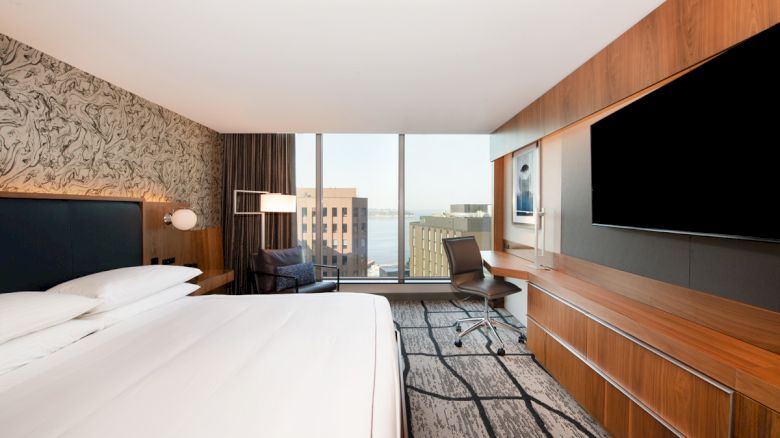 A modern hotel room with a large bed, wall-mounted TV, desk, chair, and a window view of buildings and the waterfront.