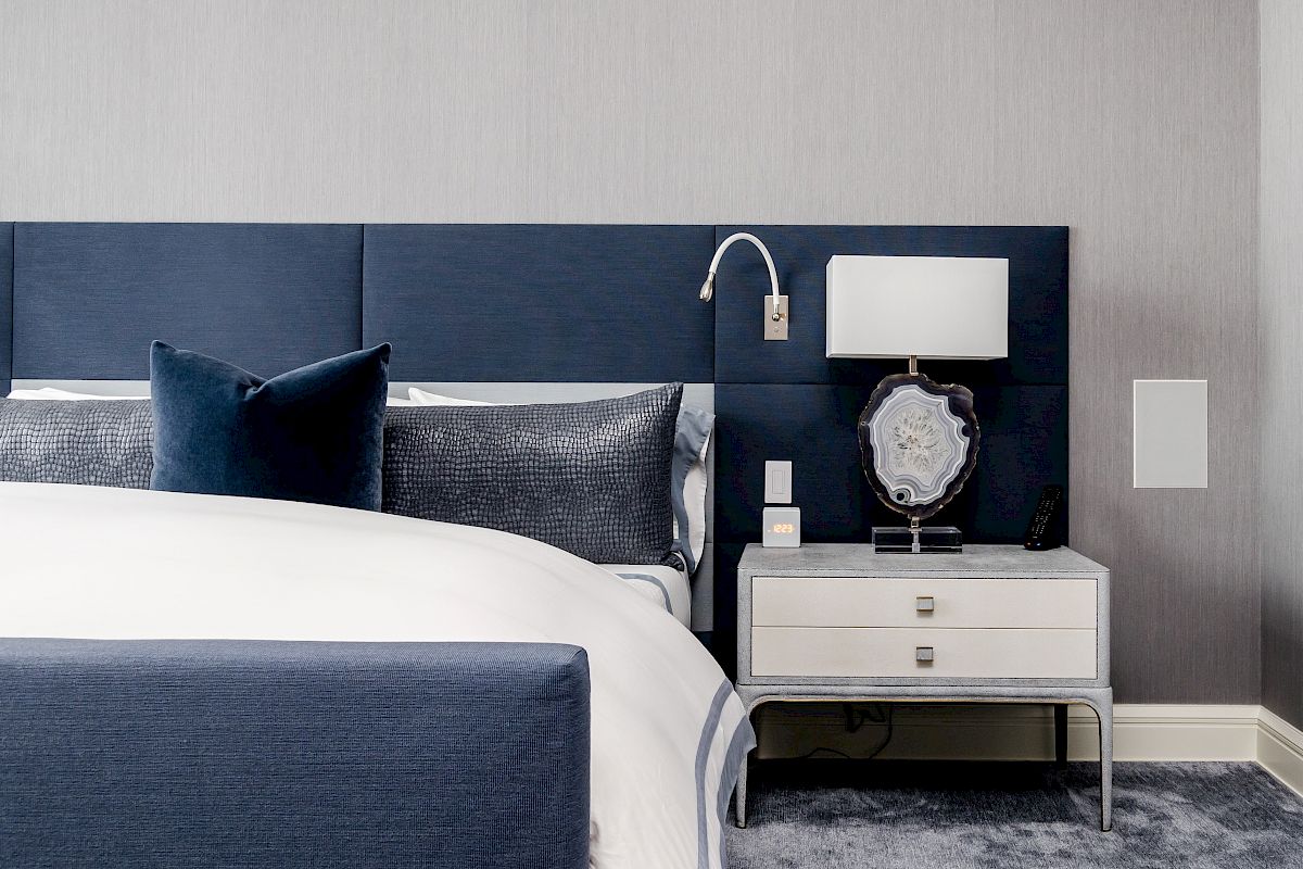 A modern bedroom features a blue upholstered bed, gray walls, a stylish nightstand, a decorative lamp, and a cozy carpeted floor.