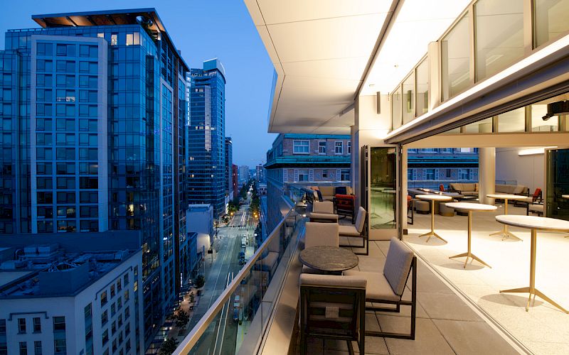 Rooftop view of a modern cityscape with tall buildings, a terrace with tables and chairs, and evening lighting.